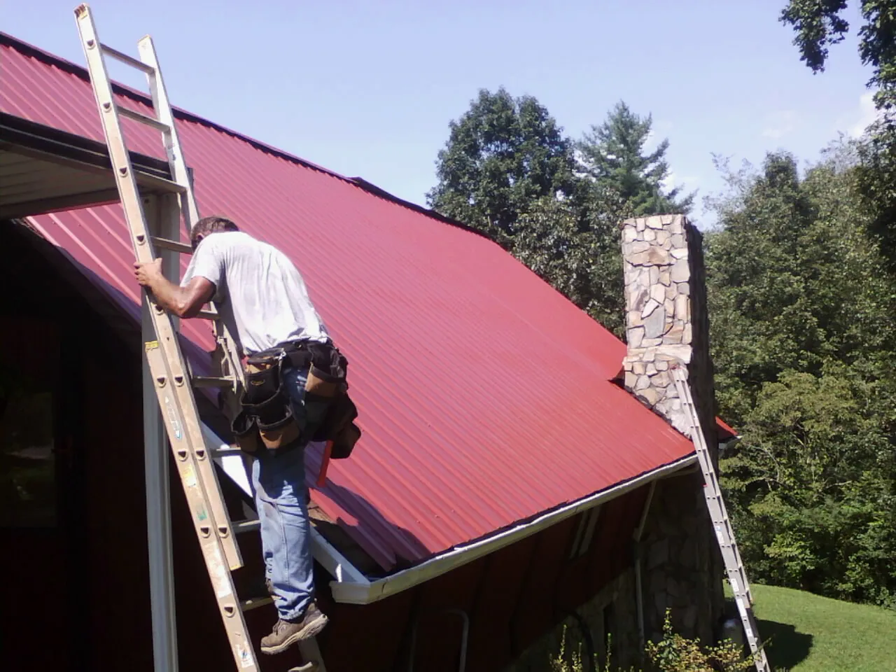 Roofer on a ladder beside a red metal roof and stone chimney — Soffit & Fascia Repair by Roofing Brothers in Toledo